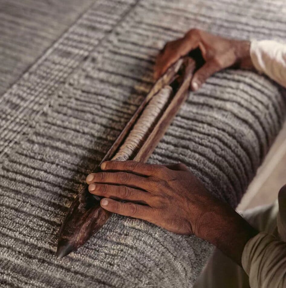 Hands weaving fabric rug with traditional wooden tool.